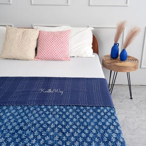 以下が含まれることがあります： 「Kantha Way」の文字が入った、ネイビーブルーと白の模様のキルトが、白い枕とピンクの模様の枕が置かれたベッドの上に置かれています。乾燥した植物が入った2つの青い花瓶が、木製のサイドテーブルに置かれています。