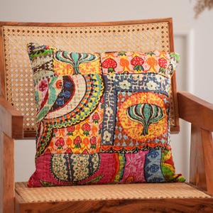 Puede incluir: Un cojín de patchwork colorido con una variedad de patrones, incluyendo diseños florales y geométricos. El cojín presenta una mezcla de colores brillantes como el rojo, amarillo, azul y rosa. Está colocado sobre una silla de madera con asiento tejido.