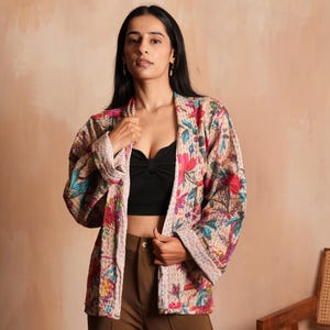 Peut inclure: Veste à imprimé floral avec un fond crème et des fleurs colorées roses, bleues et vertes. La veste a une coupe décontractée et est portée sur un haut noir et un pantalon marron. Les manches de la veste sont retroussées et les bords sont bordés de tissu crème.