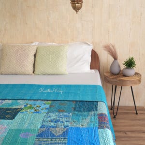 May include: A bed with a turquoise patchwork quilt and matching bed runner. The quilt features various shades of blue and green, with visible stitching. The text "Kantha Way" is on the bed runner. Decorative pillows and a small side table are also visible.