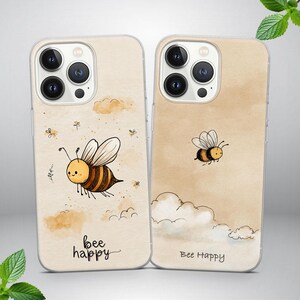 Könnte beinhalten: Zwei Handyhüllen mit beigem Aquarellhintergrund und einem Cartoon-Bienen-Design. Eine Hülle zeigt eine Biene mit dem Text "bee happy", die andere eine fliegende Biene mit demselben Text. Die Hüllen haben einen transparenten Rand.