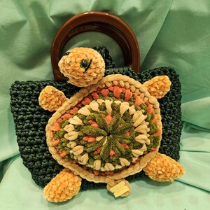 Puede incluir: Bolso de crochet verde oscuro hecho a mano con un diseño de tortuga. La tortuga tiene un caparazón marrón, naranja y verde, con apéndices a juego en forma de aleta. La cabeza de la tortuga es marrón con un ojo verde. El bolso tiene un asa de madera marrón.