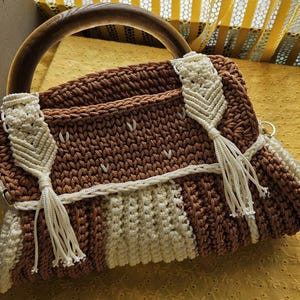 May include: A brown and cream-colored macrame handbag with a wooden handle. The bag features a woven design with cream-colored accents and tassels. The bag is rectangular and has a curved wooden handle.