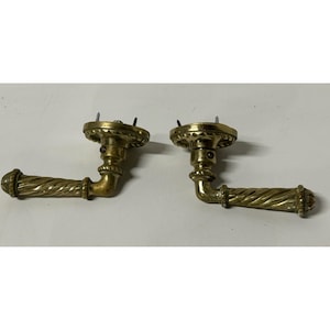 May include: Two antique brass door handles. Each handle features a decorative, twisted design on the grip and a round, ornate base. The handles are attached with screws, suggesting easy installation. Ideal for vintage home decor.
