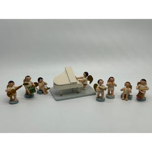 May include: A collection of small, wooden angel figurines playing musical instruments. The set includes a grand piano, guitar, drums, and other instruments. The angels have gold wings and are painted in neutral colors.