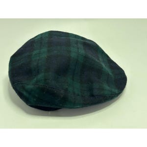May include: A flat cap featuring a dark green and navy blue plaid pattern. The hat has a rounded, low-profile crown and a short, stiff brim at the front. The fabric displays a soft, textured appearance with a classic intersecting check design.