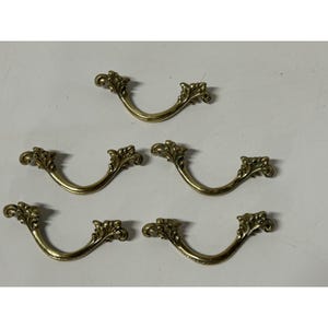 May include: Set of five brass-toned drawer pulls. Each pull has a curved design with ornate floral detailing at each end. The pulls are arranged on a white surface, showcasing their decorative style.