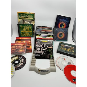 May include: A collection of computer games and CDs, including "Where in the World is Carmen Sandiego?", "Warcraft II: Tides of Darkness", and "Pinball". The items are arranged on a white surface with a Gateway 2000 CD holder.