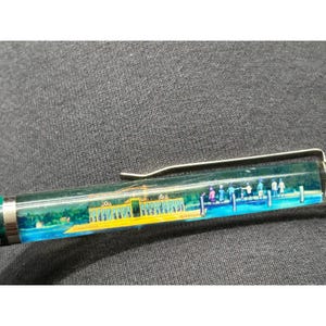 May include: A green pen with a clear barrel featuring a colorful illustration of a yellow building and people on a pier. The pen has a silver clip and a green cap.