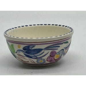 May include: A small, off-white ceramic bowl with a hand-painted design. The bowl features a blue bird, purple flowers, and green leaves. The rim has a dotted blue pattern. The bowl is likely for serving or display.