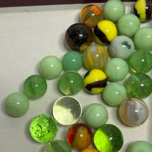 May include: A collection of colorful glass marbles in a variety of colors, including green, yellow, orange, and black. The marbles are arranged in a box.