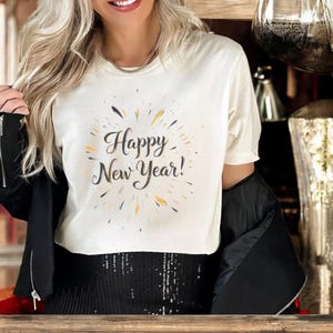 Puede incluir: Camiseta color crema con la frase "Happy New Year!" en fuente cursiva, rodeada de acentos coloridos. La camiseta se combina con una chaqueta negra y una falda con lentejuelas, creando un look festivo.