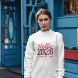 Puede incluir: Sudadera blanca con el texto "Hello 2026" en letras rosas y marrones brillantes. La frase "Let's Try This Again" está debajo. Una persona lleva la sudadera delante de un edificio azul.