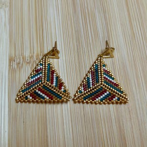 Puede incluir: Un par de pendientes triangulares dorados. Los pendientes están adornados con un patrón de cuentas en color turquesa, rojo, blanco y dorado. Los pendientes se exhiben sobre una superficie de madera.