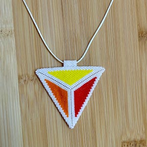 Puede incluir: Un collar con colgante hecho a mano. El colgante es un triángulo blanco con secciones de cuentas amarillas, naranjas y rojas. El collar tiene un cordón blanco. El colgante mide aproximadamente 5 cm de ancho.