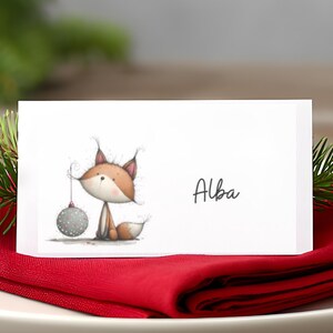 Puede incluir: Un montaje de mesa navideño con un plato blanco, una servilleta roja y una tarjeta con una ilustración de un zorro sosteniendo una bola de Navidad. Una vela roja encendida y ramitas de pino completan el ambiente festivo.