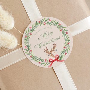 May include: A Christmas gift wrapped in brown paper, with a cream ribbon and a round tag. The tag features a watercolour wreath, berries, and a reindeer, with the words "Merry Christmas". Two fluffy, dried, cream-coloured flowers are attached.