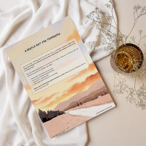 Op de afbeelding: Een rechthoekige papieren print getiteld A GENTLE GIFT FOR TOMORROW met een landschapsillustratie met oranje en roze wolken boven heuvels. De tekst bevat suggesties voor zelfzorg, inclusief de zin A little kindness, just for you.