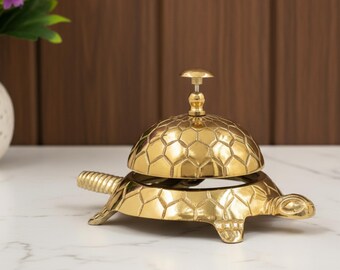 May include: A decorative brass turtle-shaped bell. The bell features a hexagonal pattern on its dome and a small handle. The turtle's body forms the base, with its head and tail extending outwards. The bell is made of brass.