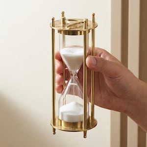 Puede incluir: Un reloj de arena con marco de latón y cámaras de vidrio transparente llenas de arena blanca. El reloj de arena se sostiene en una mano, mostrando su intrincado diseño y el flujo del tiempo. La pieza decorativa tiene una estética vintage.