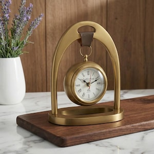 Peut inclure: Une horloge de bureau dorée en forme d'arche avec un cadran rond et des chiffres romains. L'horloge est suspendue à une lanière en cuir dans l'arche. La base est un rectangle en bois foncé. Le cadran de l'horloge indique "Franklin & Murphy."
