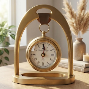May include: A gold-colored desk clock with a round face and Roman numerals. The clock is suspended within a gold arch and base. The clock face reads "Franklin Murphy Oxford". A brown leather strap connects the clock to the arch.