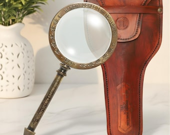 May include: A vintage brass magnifying glass with a clear lens, resting next to a brown leather case. The magnifying glass is engraved with "HENRY HUGHES SON LTD. LONDON - 1941". The leather case has an anchor emblem.