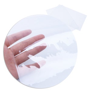 May include: Clear plastic sheet, rolled up and held by a hand. The sheet is translucent and shows a slight ripple pattern.
