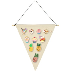 May include: A white triangular canvas with a wooden dowel at the top and a black string hanging from the top corners. The canvas is decorated with 12 colourful enamel pins featuring various desserts and pineapples.