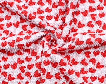 May include: A textured fabric featuring a repeating pattern of small, solid red hearts printed on a white background. The material is gathered in the center, creating folds and shadows that highlight the puckered, seersucker-like surface texture.