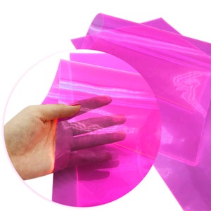 May include: A sheet of translucent pink iridescent vinyl. The vinyl is folded in half and a hand is holding the sheet up.