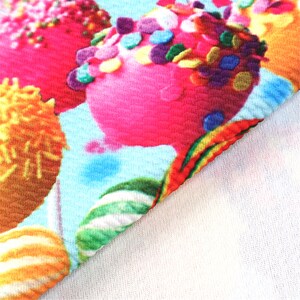 May include: A close-up of a colorful fabric with a repeating pattern of pink, yellow, and green candy-like shapes covered in sprinkles. The fabric has a textured, waffle-like surface.