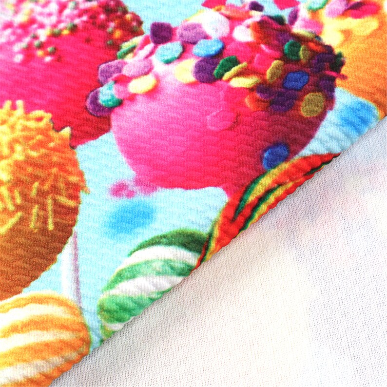 May include: A close-up of a colorful fabric with a repeating pattern of pink, yellow, and green candy-like shapes covered in sprinkles. The fabric has a textured, waffle-like surface.