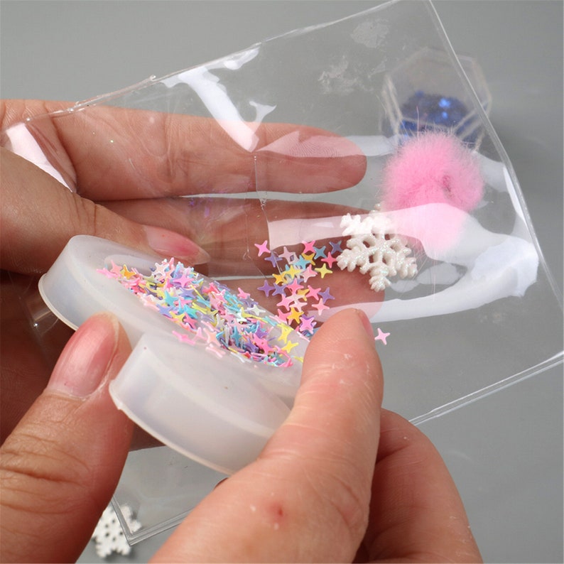 May include: A clear plastic bag filled with colorful star-shaped sprinkles, a white snowflake, and a pink pom-pom. The bag is being held over a white silicone mold shaped like a heart.