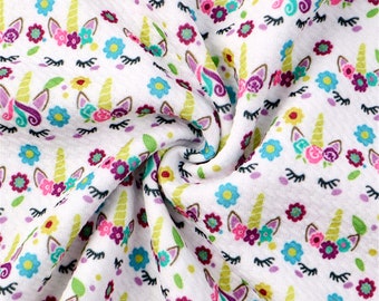 May include: White fabric with a repeating pattern of unicorn faces with flowers and eyelashes. The fabric is folded and twisted.