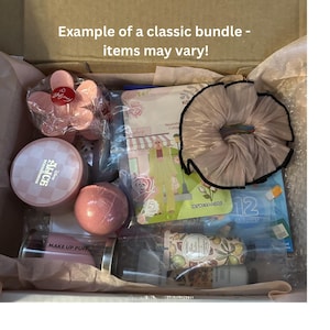 May include: A box filled with beauty products, including a pink flower-shaped container, a round pink compact, a pink bath bomb, and a scrunchie. The box also contains a tube of lotion, a face mask, and a makeup puff. Text on the box reads "Example of a classic bundle - items may vary!"