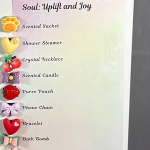 May include: A white background with the text "Soul: Uplift and Joy" and a list of items including a scented sachet, shower steamer, crystal necklace, scented candle, purse pouch, phone chain, bracelet, bath bomb, and pocket mirror. The image is decorated with flower-shaped embellishments.