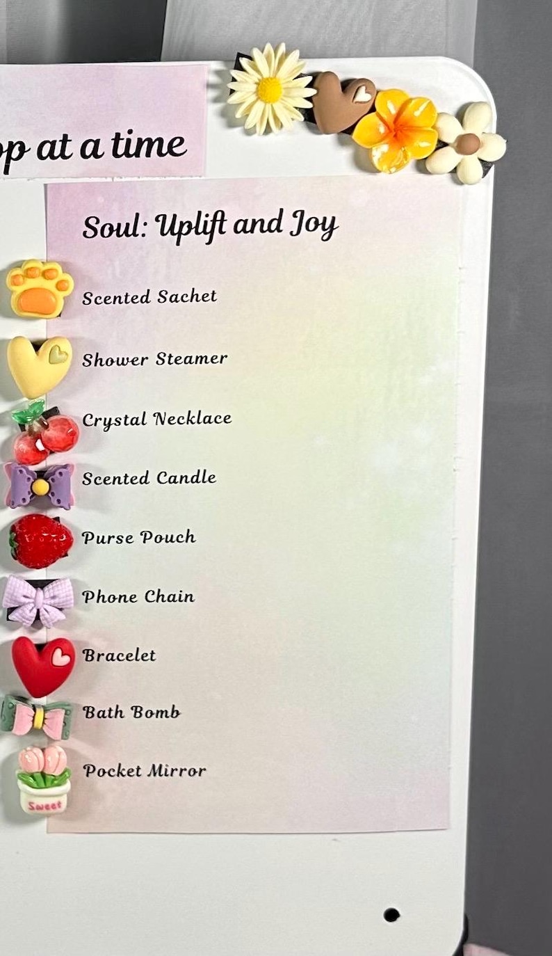 May include: A white background with the text "Soul: Uplift and Joy" and a list of items including a scented sachet, shower steamer, crystal necklace, scented candle, purse pouch, phone chain, bracelet, bath bomb, and pocket mirror. The image is decorated with flower-shaped embellishments.