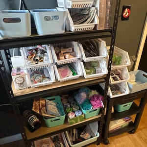 May include: A multi-tiered storage rack filled with various items. The top shelves hold light blue bins and white plastic baskets. The lower shelves contain more bins and baskets with a variety of products, including jewelry, bath products, and stationery.