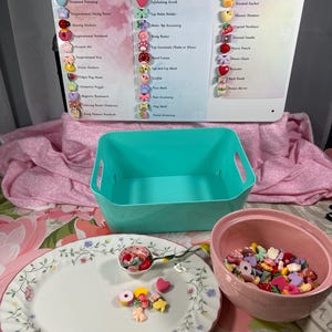 May include: A display featuring a sign with the text "Serenity Scoops" and product categories. A teal bin, a pink bowl, and a floral plate with a spoon are in the foreground. The plate and spoon hold colorful, decorative embellishments.
