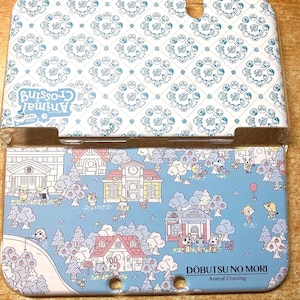 Puede incluir: Una funda azul y blanca para Nintendo 3DS. La tapa superior presenta un diseño estampado, mientras que la parte inferior muestra una escena de pueblo con animales de dibujos animados. Incluye el texto "Animal Crossing" y "Dōbutsu no Mori".