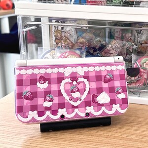 May include: A pink and white Nintendo 3DS case featuring a gingham pattern with strawberries and whipped cream accents. The case has a decorative lace trim and a heart-shaped strawberry design in the center.