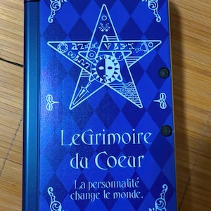 May include: A blue handheld gaming device with a cover featuring a star design and the text "Le Grimoire du Coeur" and "La personnalité change le monde." The cover has a diamond pattern and decorative corner elements.