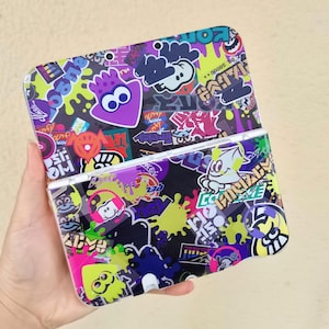 Puede incluir: Una funda para consola de juegos portátil con un diseño colorido. La funda está cubierta con personajes de dibujos animados, salpicaduras y texto japonés en tonos morados, verdes, amarillos y rosas. La funda está abierta, mostrando la pantalla y los botones.