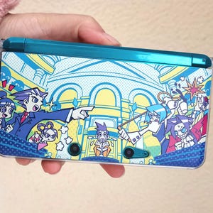Op de afbeelding: Een teal Nintendo 3DS handheld gameconsole met een kleurrijke cartoonafbeelding. De afbeelding toont meerdere personages in een rechtszaal met tekstballonnen en actielijnen. De console wordt in een hand gehouden.