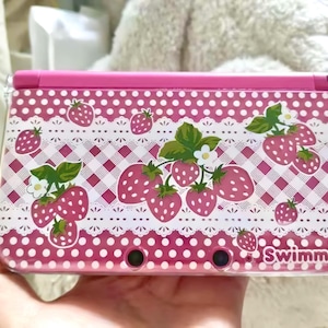 Könnte beinhalten: Rosa Nintendo 3DS-Hülle mit Erdbeermuster. Die Hülle hat ein rosa-weißes Karomuster, einen weißen Spitzensaum und Illustrationen von Erdbeeren mit grünen Blättern und weißen Blüten. Das Wort "Swimmer" ist unten rechts aufgedruckt.
