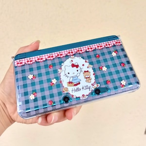 May include: A blue and purple Nintendo 3DS case featuring a Hello Kitty design. The case has a gingham pattern with cherries, flowers, and strawberries. The Hello Kitty graphic is in a white oval with the words "Hello Kitty".