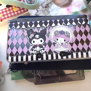 Könnte beinhalten: Eine schwarze und lila Nintendo 3DS-Hülle mit Zeichentrickfiguren. Die Hülle hat ein Harlekinmuster in Schwarz und Lila mit einem schwarzen Rand. Die Figuren befinden sich in der Mitte.