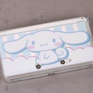 May include: A white Nintendo 3DS handheld game console with a custom cover. The cover features a cartoon character on a light blue and white striped background. The cartoon character is white with pink accents and large ears.