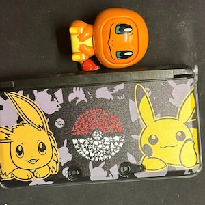 May include: A black Nintendo 3DS console with a colorful Pokemon design. The console features illustrations of Pikachu and Eevee, along with a Poke Ball graphic. An orange Charmander figure is on top of the console.